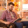 young man sits on his couch reading a book next to his sleeping dog